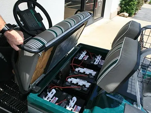 Golf cart seat lifted to reveal battery compartment outside a building in sunny weather