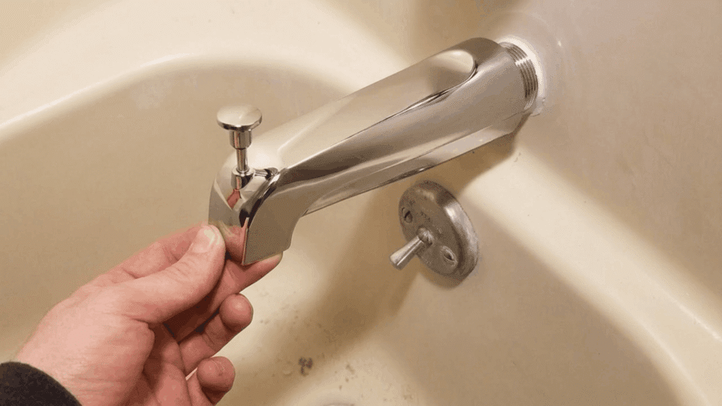hand holding a chrome tub spout with a diverter near the bathtub wall showing pipe connection during the installation process