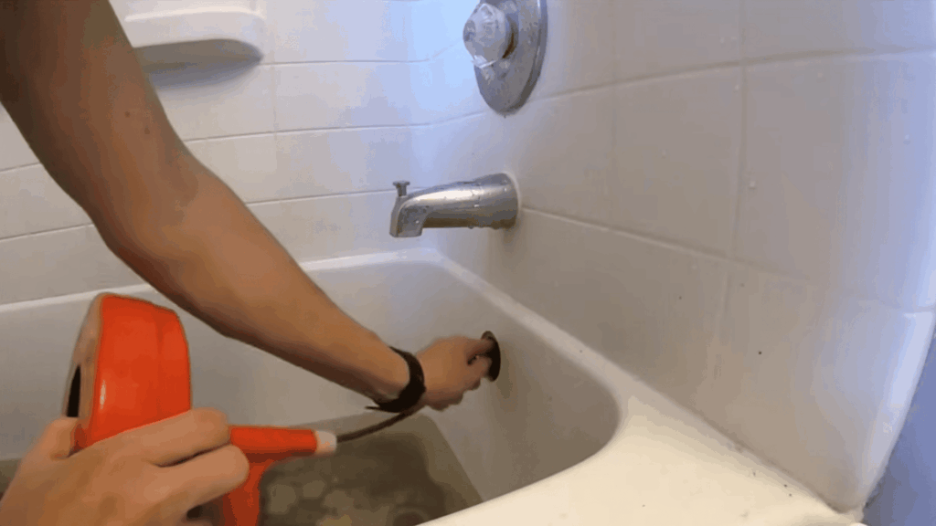 hand using drain snake tool in bathtub overflow opening showing how to unclog a bathtub drain step