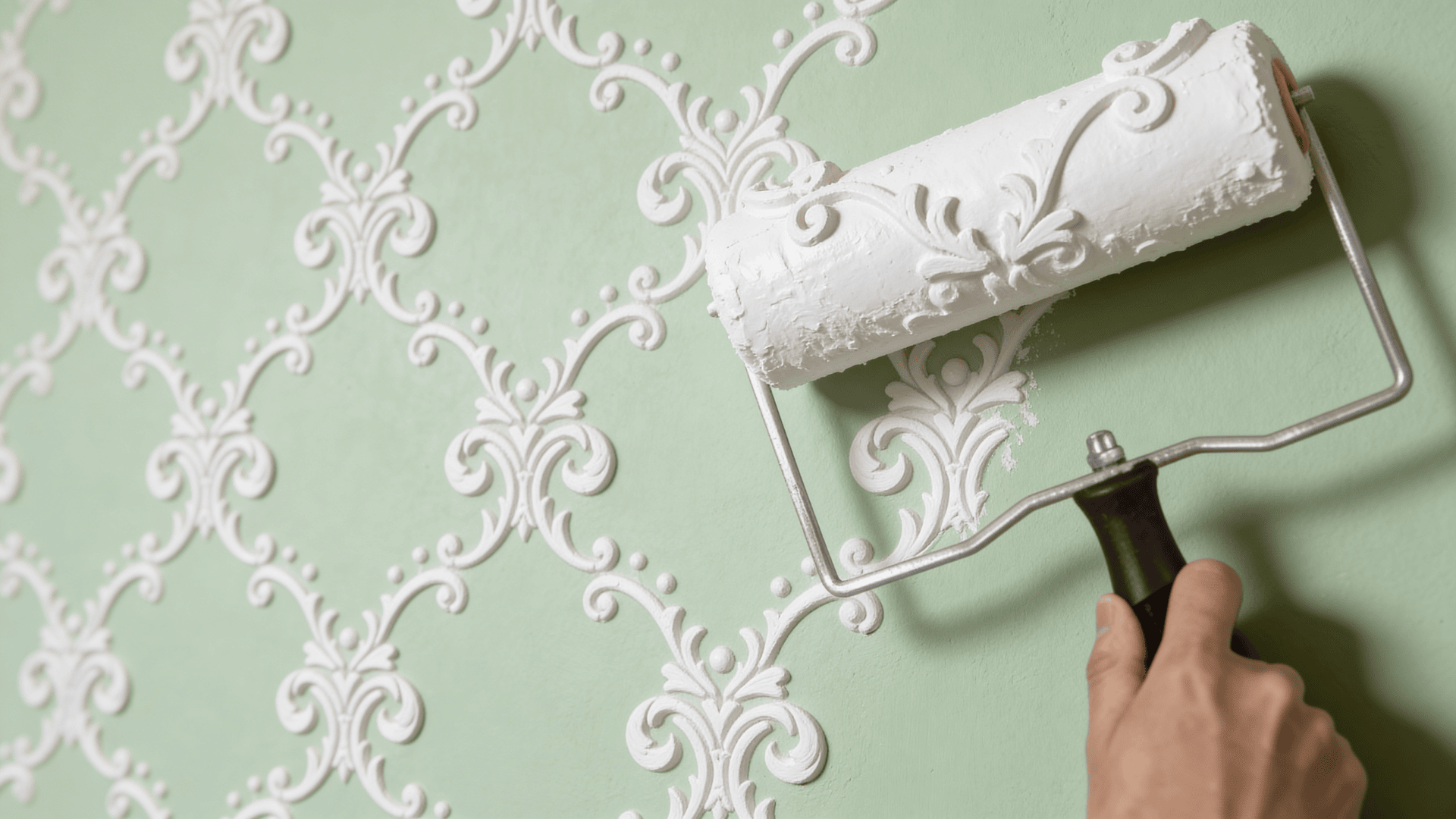 hand using paint roller to apply white decorative pattern over green wall creating ornate textured design