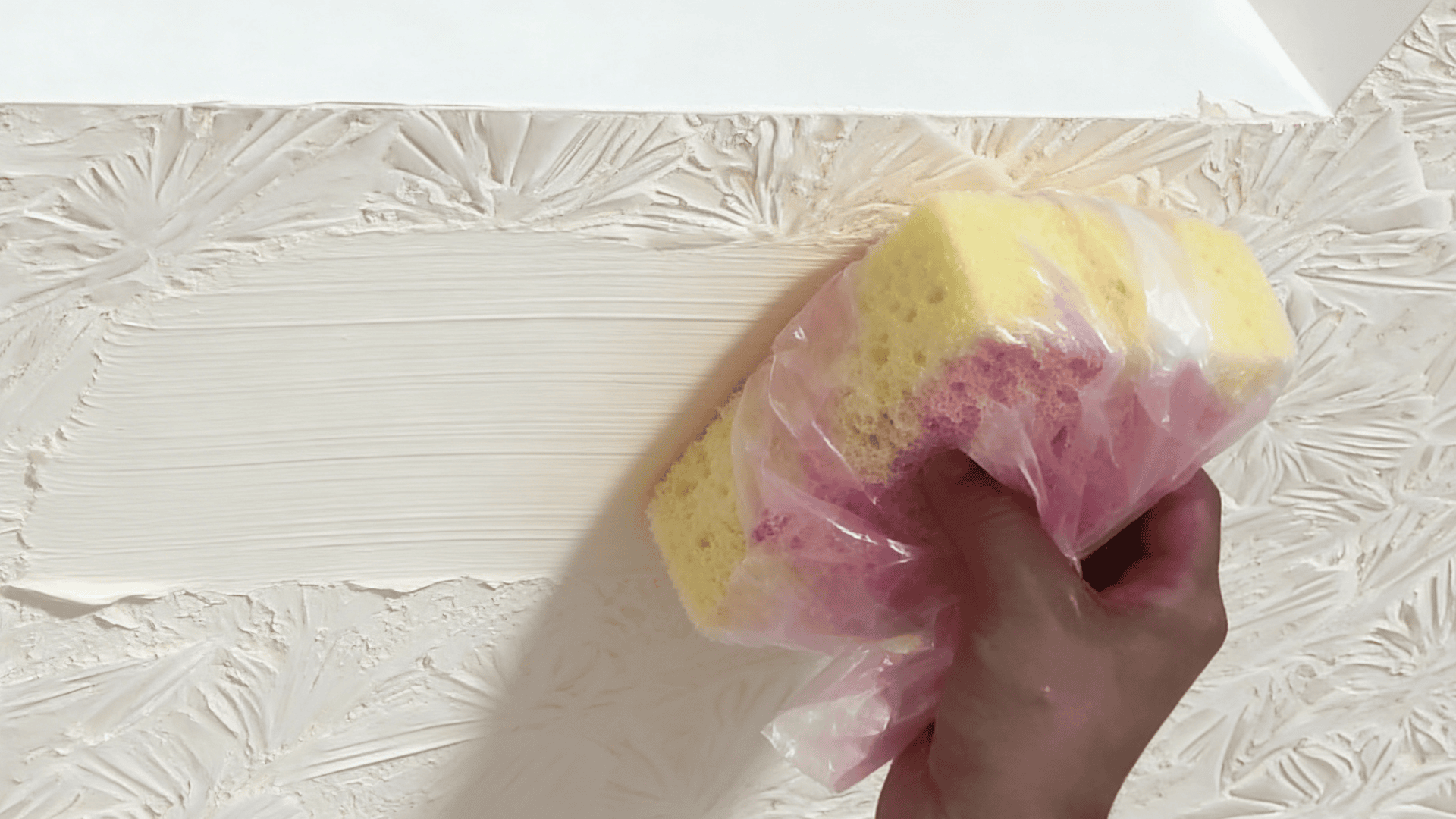 hand using sponge wrapped in plastic applying paint over textured ceiling surface with floral patterns near light opening