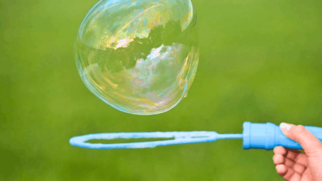 homemade bubble solution hand holding blue bubble wand blowing large soap bubble floating in air against green outdoor background