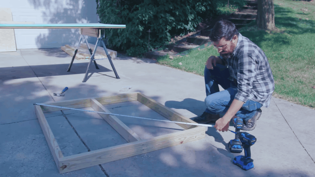 man measuring wooden base frame on ground with drill nearby