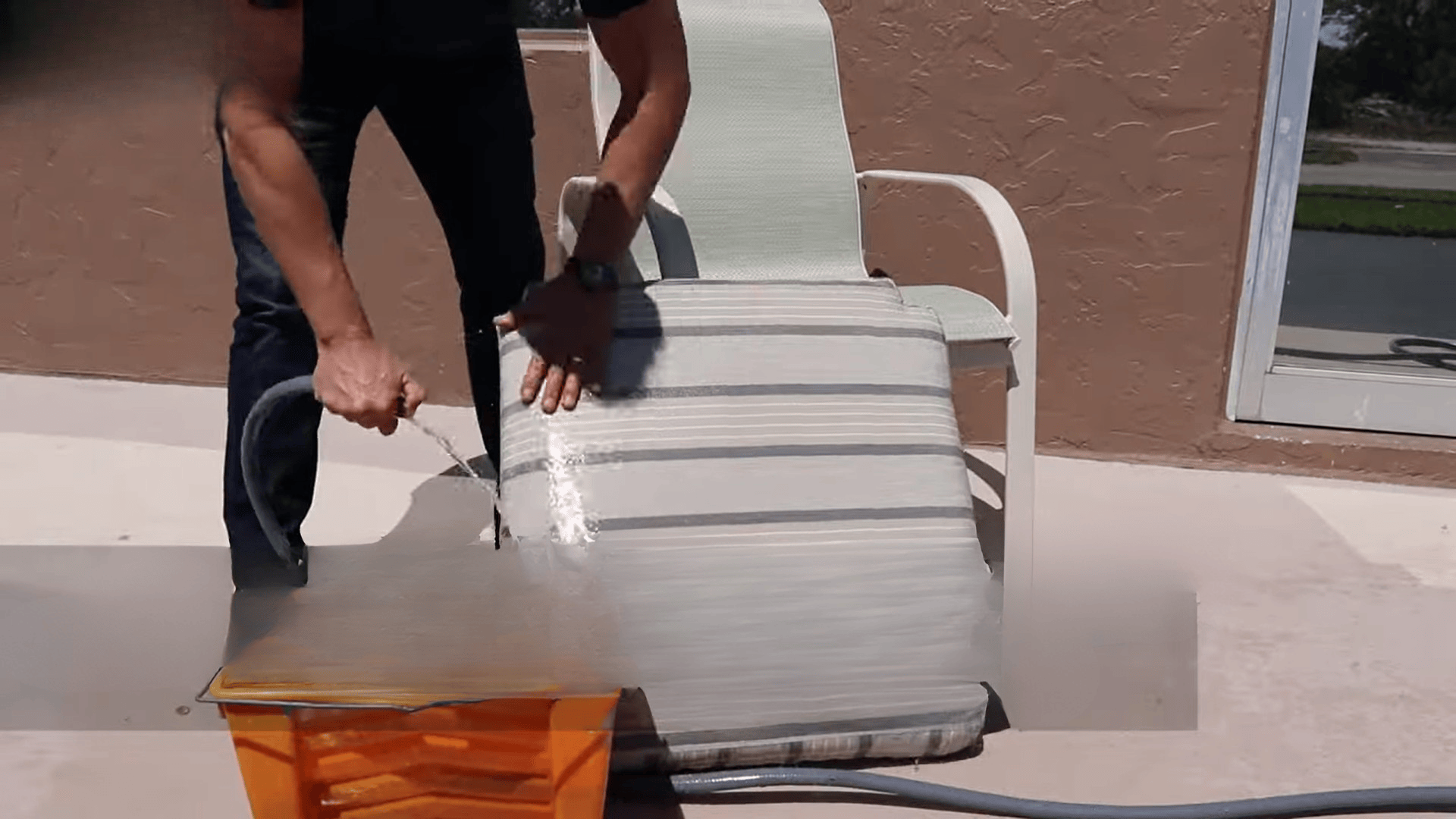 person holding bucket with soapy water near outdoor cushion demonstrating how to clean sunbrella cushions using washing tools and hose (1)