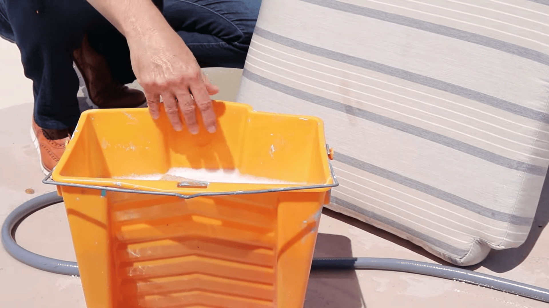 person holding bucket with soapy water near outdoor cushion demonstrating how to clean sunbrella cushions using washing tools and hose