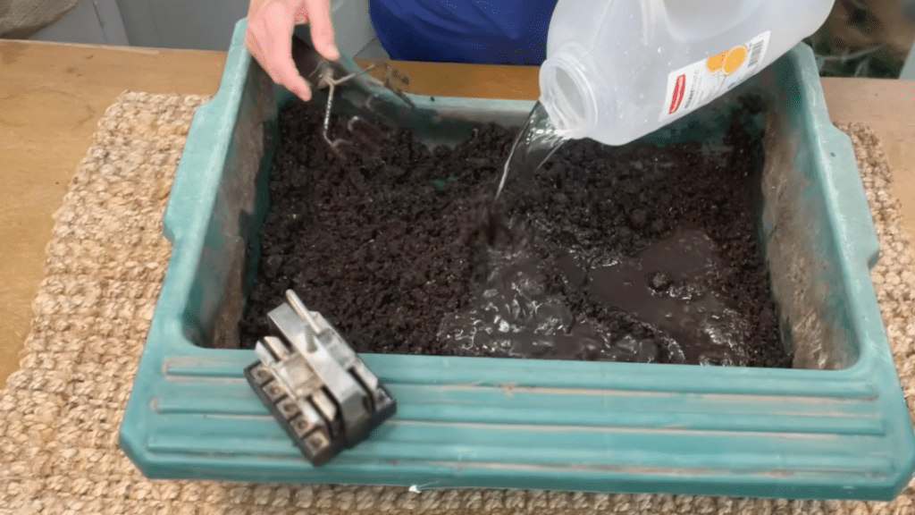 person pouring water into soil mix in tray while using hand tool to prepare moist soil for strong seed growth