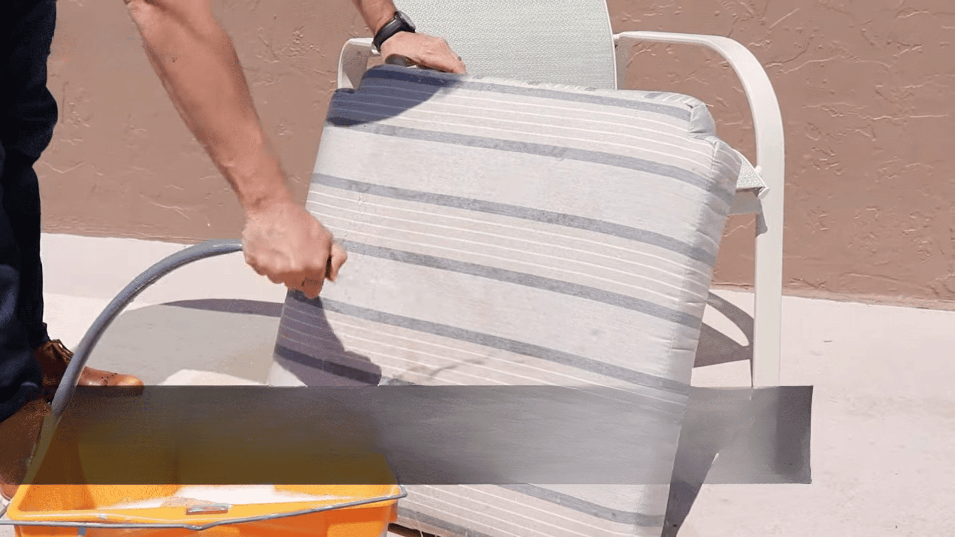 person rinsing outdoor cushion with water hose near bucket demonstrating how to clean sunbrella cushions by removing dirt and debris (1)