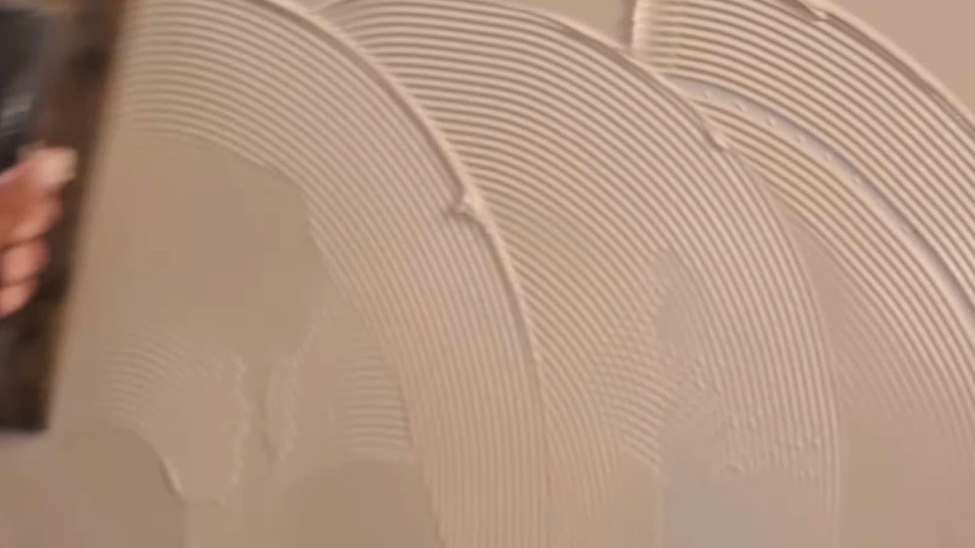 person using notched trowel to create circular patterns on wet plaster forming decorative textured wall finish during interior wall design process