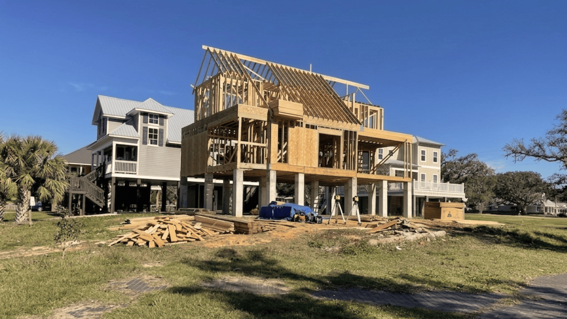 raised wooden house frame under construction on stilts with roof trusses visible beside finished homes and open yard