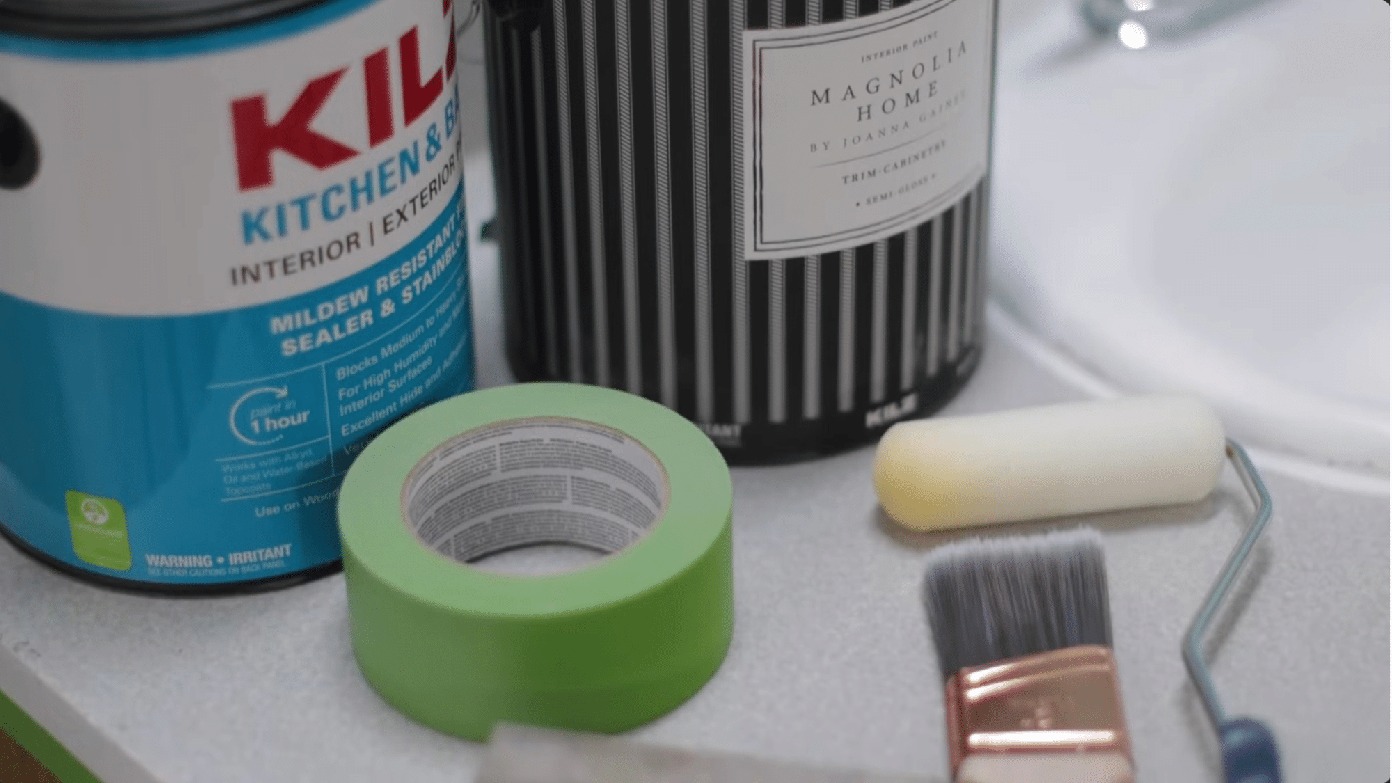supplies for painting bathroom cabinets including primer paint roller brush and painter tape arranged on countertop