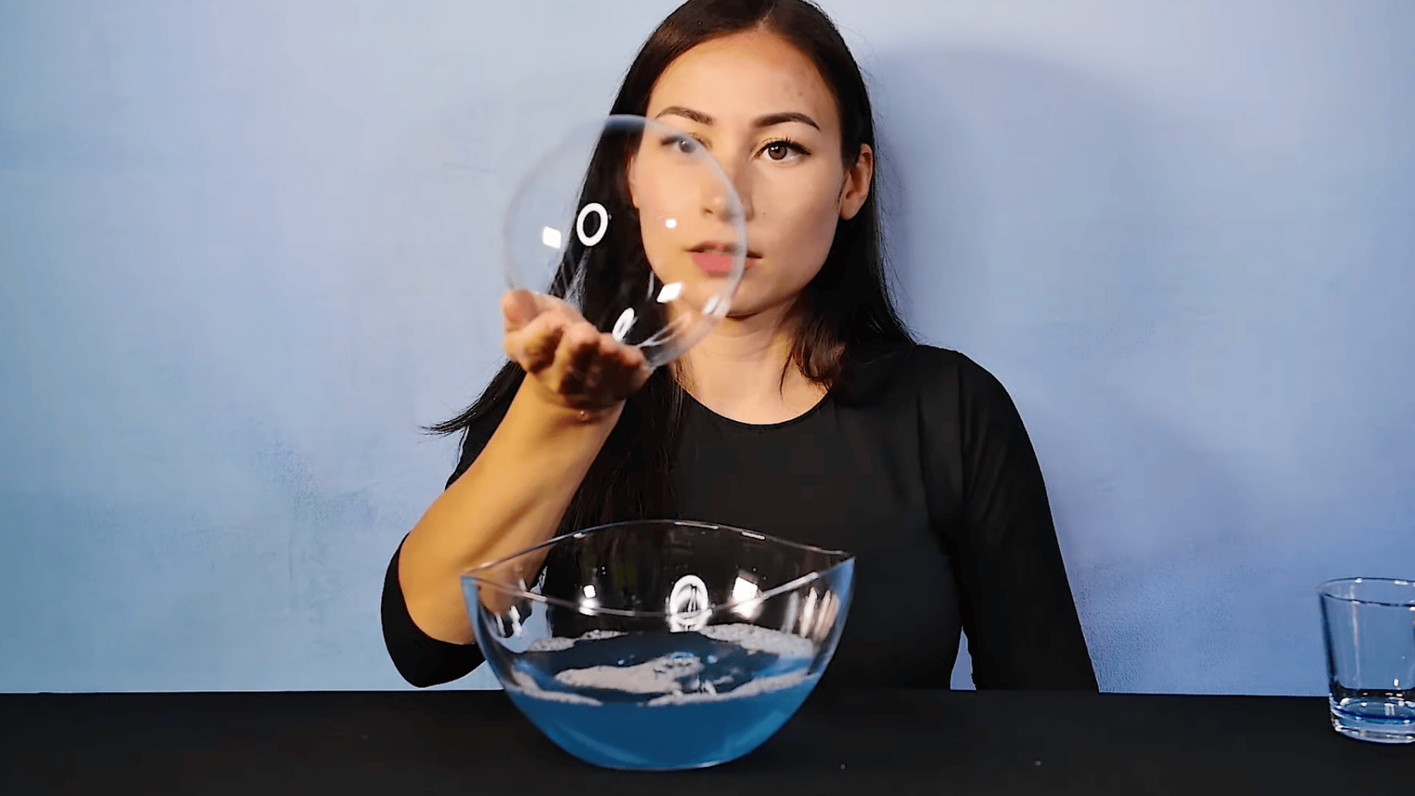 woman holding large soap bubble above bowl with blue liquid showing experiment result against plain background