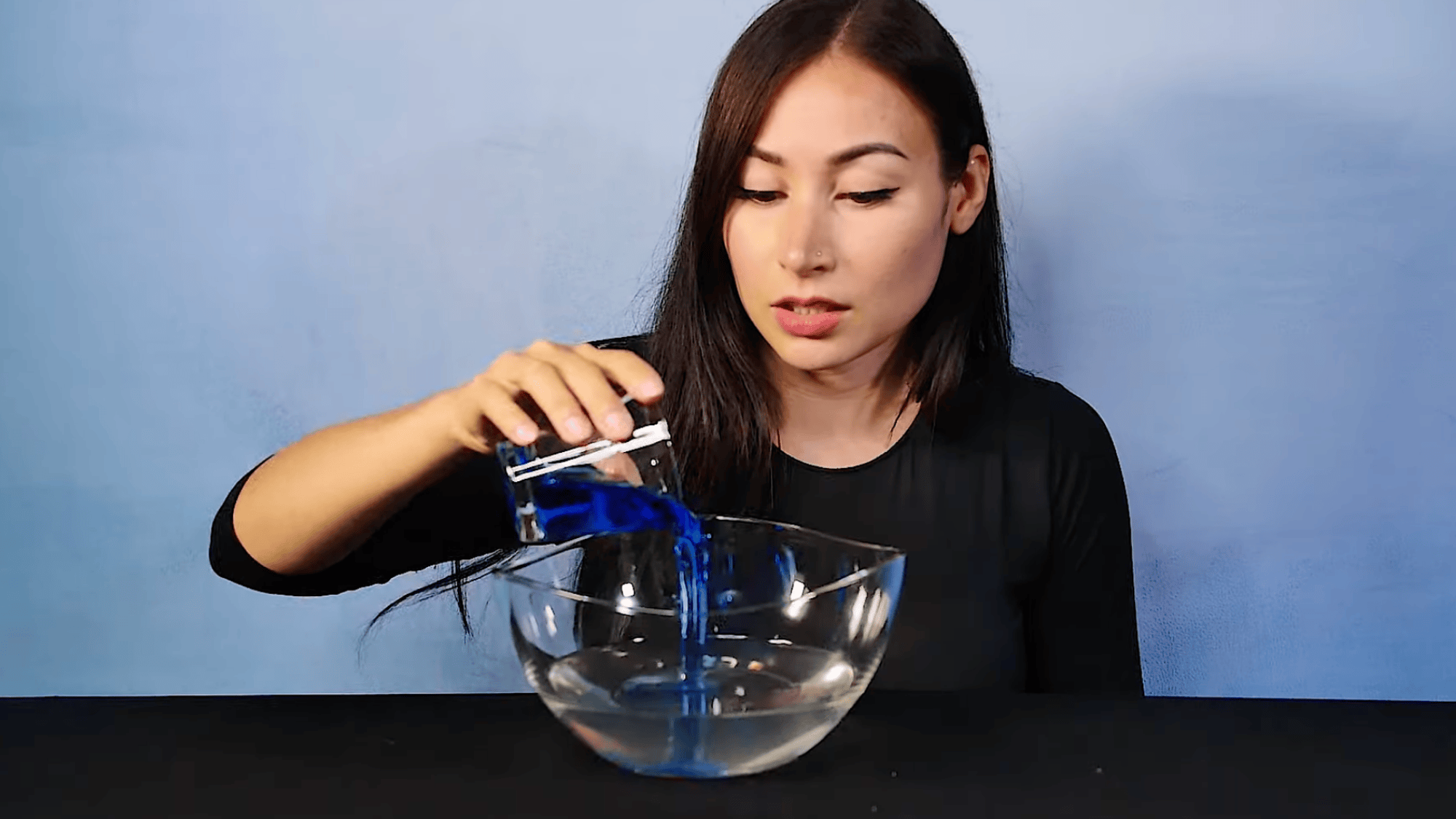 woman pouring blue liquid into glass bowl mixing with water creating colored solution on table