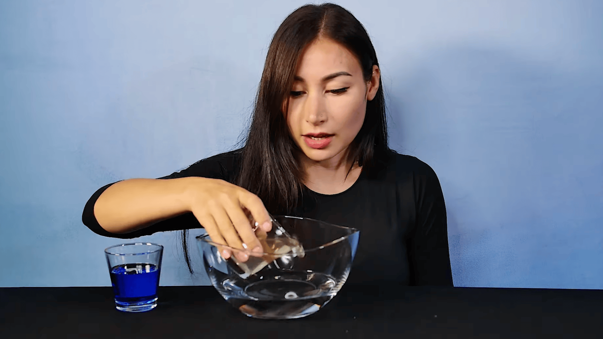 woman pouring clear liquid from small glass into empty bowl on table with blue drink beside her