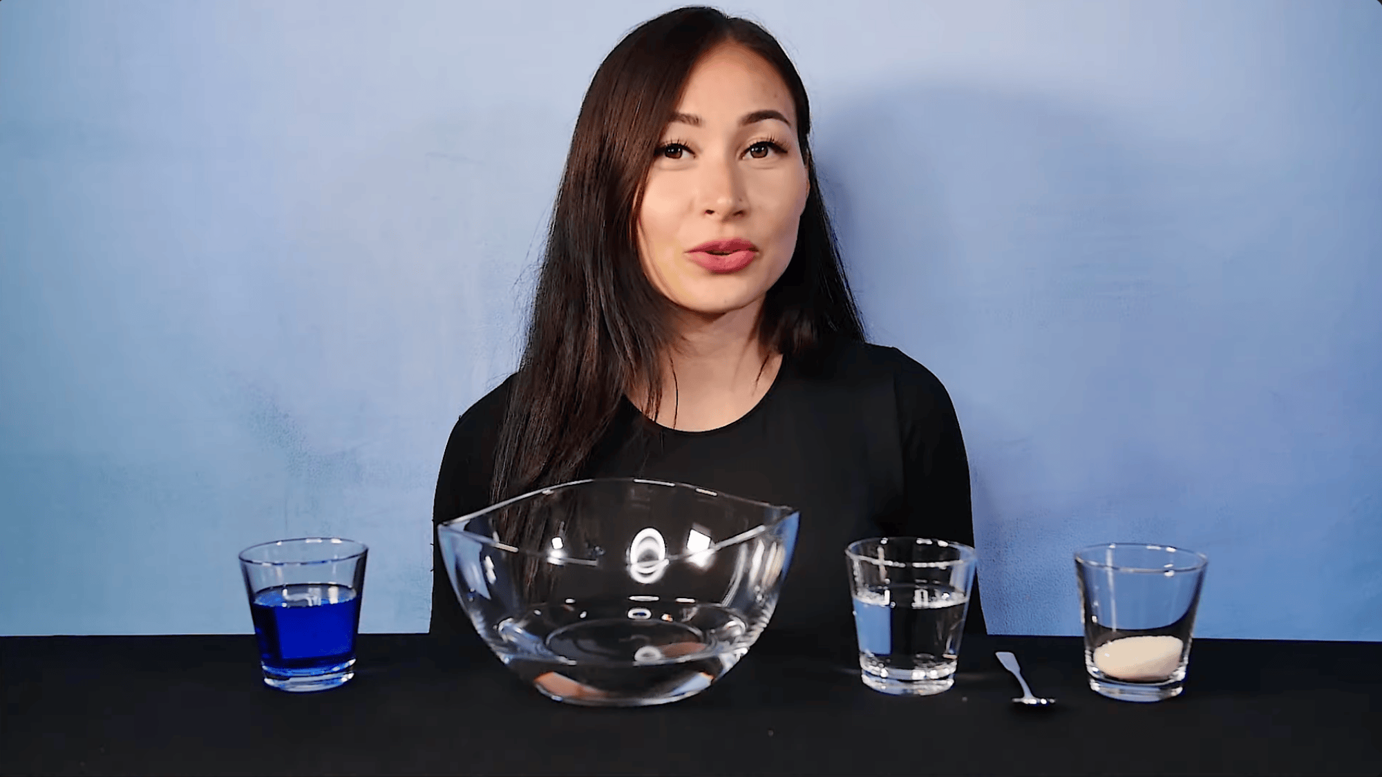 woman smiling behind table with bowl and small glasses of blue liquid water and powder showing experiment setup