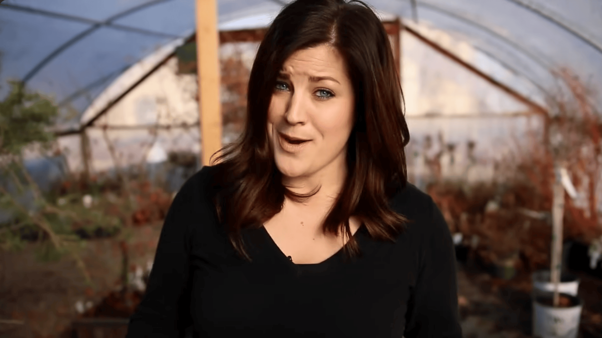 woman standing in a greenhouse with plants in background speaking to camera about succulent care and propagation