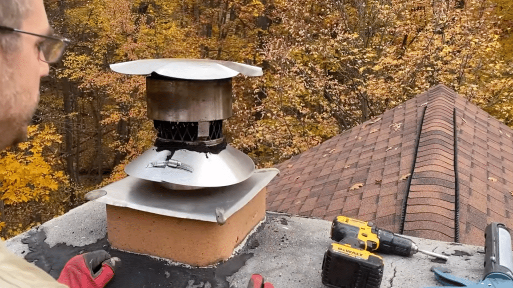 Person inspecting a metal chimney cap on a rooftop, with a drill nearby and autumn trees in the background.