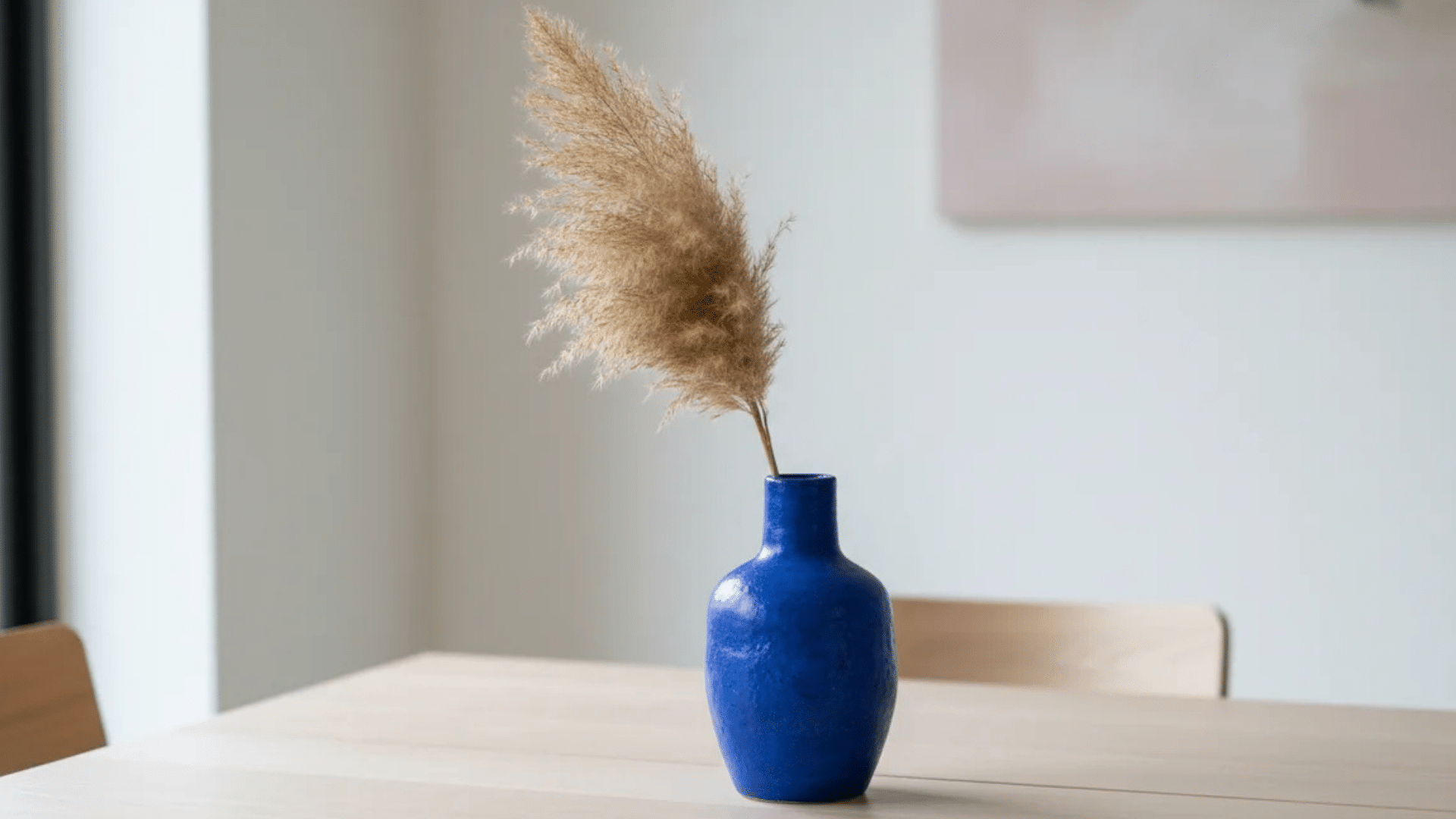 a blue vase with dried grass placed on a wooden table in a minimal room with soft natural light from window (1)