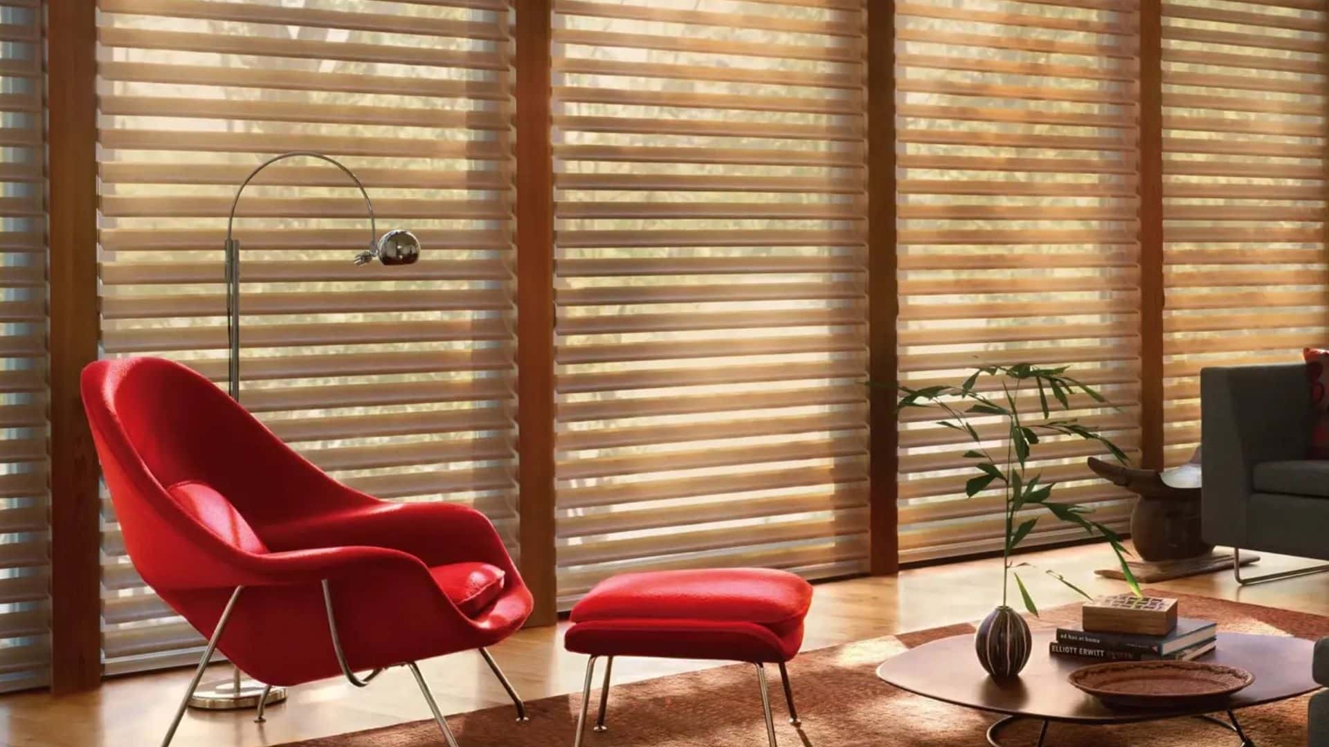a bright living room with red lounge chair and matching ottoman beside wooden blinds filtering warm sunlight across floor