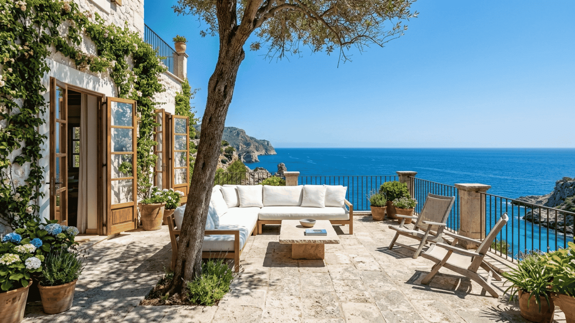 a coastal terrace with sofa chairs and table overlooking blue ocean beside a house with open doors and potted plants under clear sky