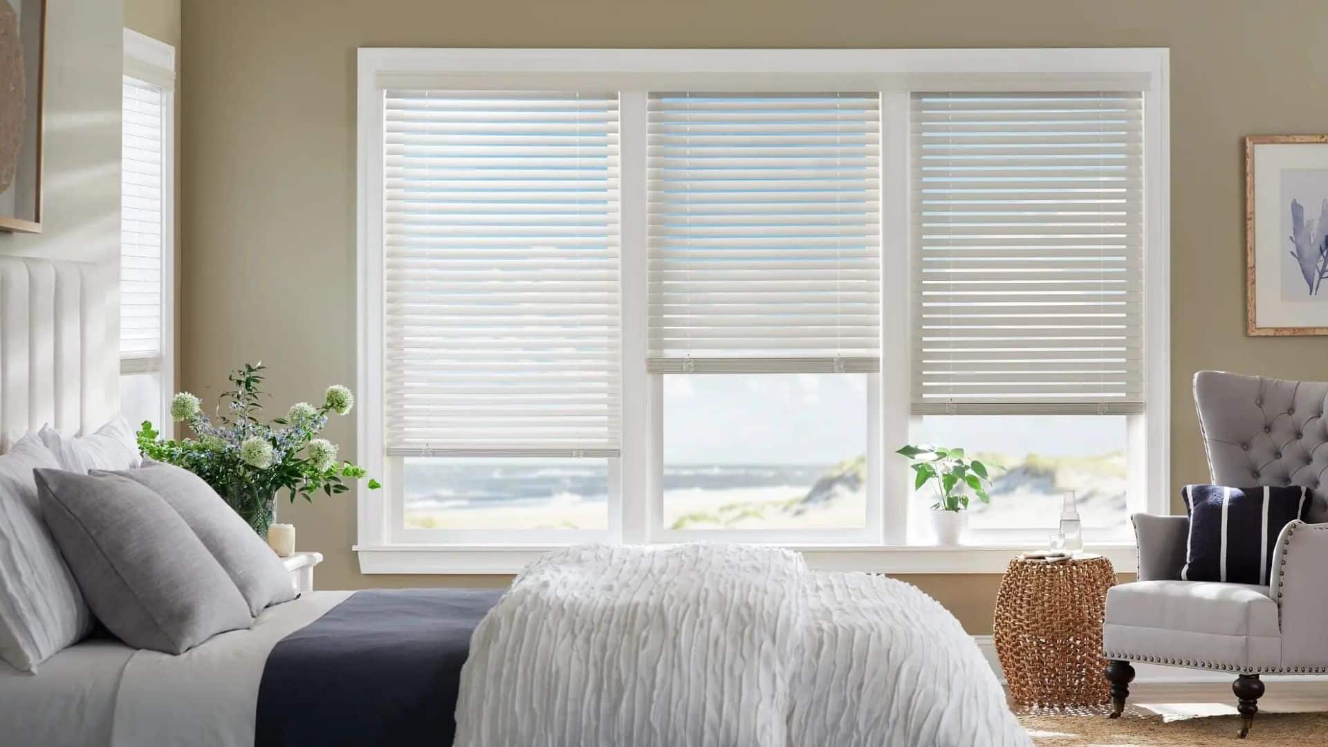a cozy bedroom with three windows fitted with blinds overlooking a beach with bed chair and plants in soft neutral tones