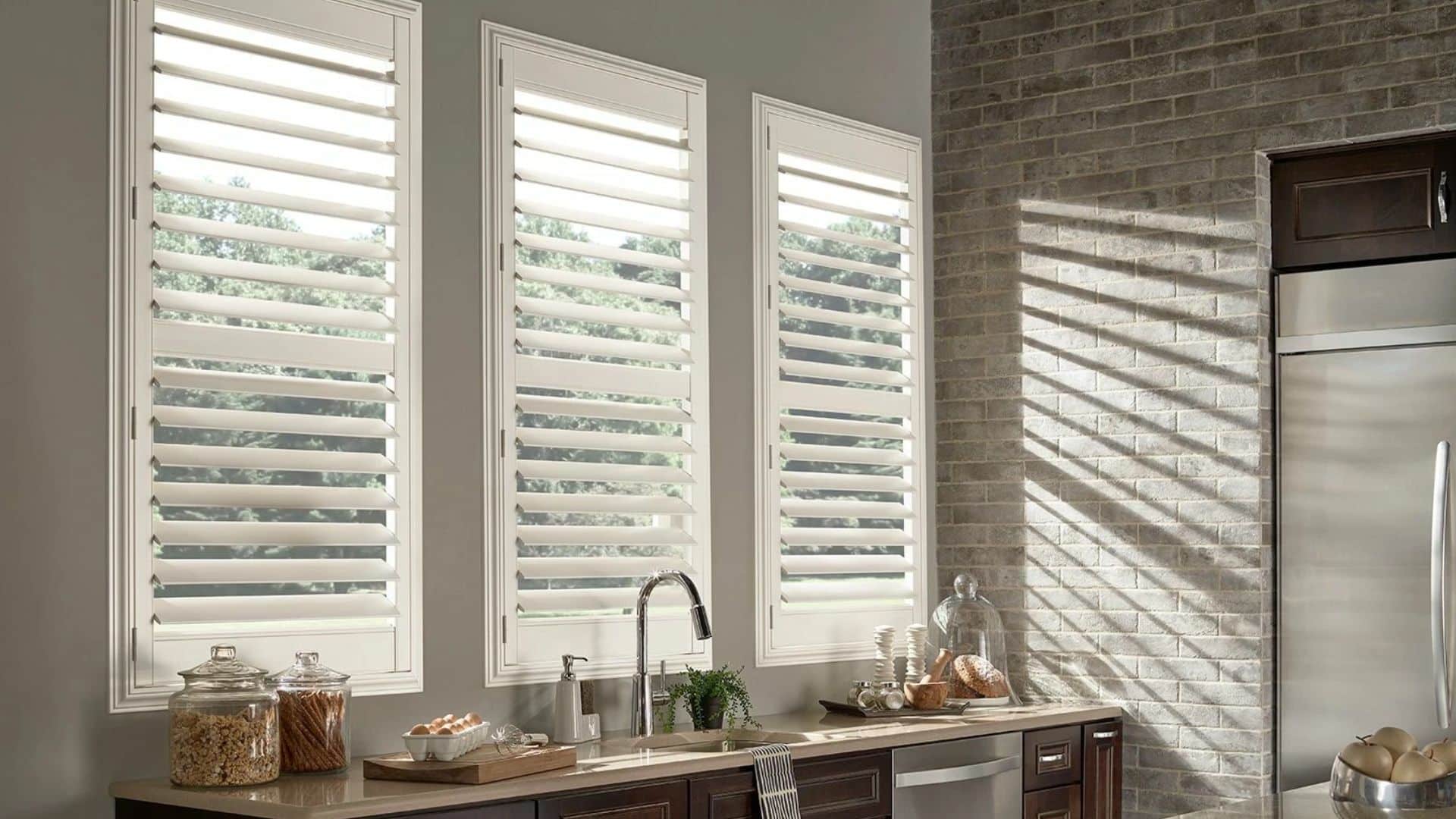 a kitchen with white plantation shutters over tall windows casting shadows on a brick wall above a sink with jars and countertop items