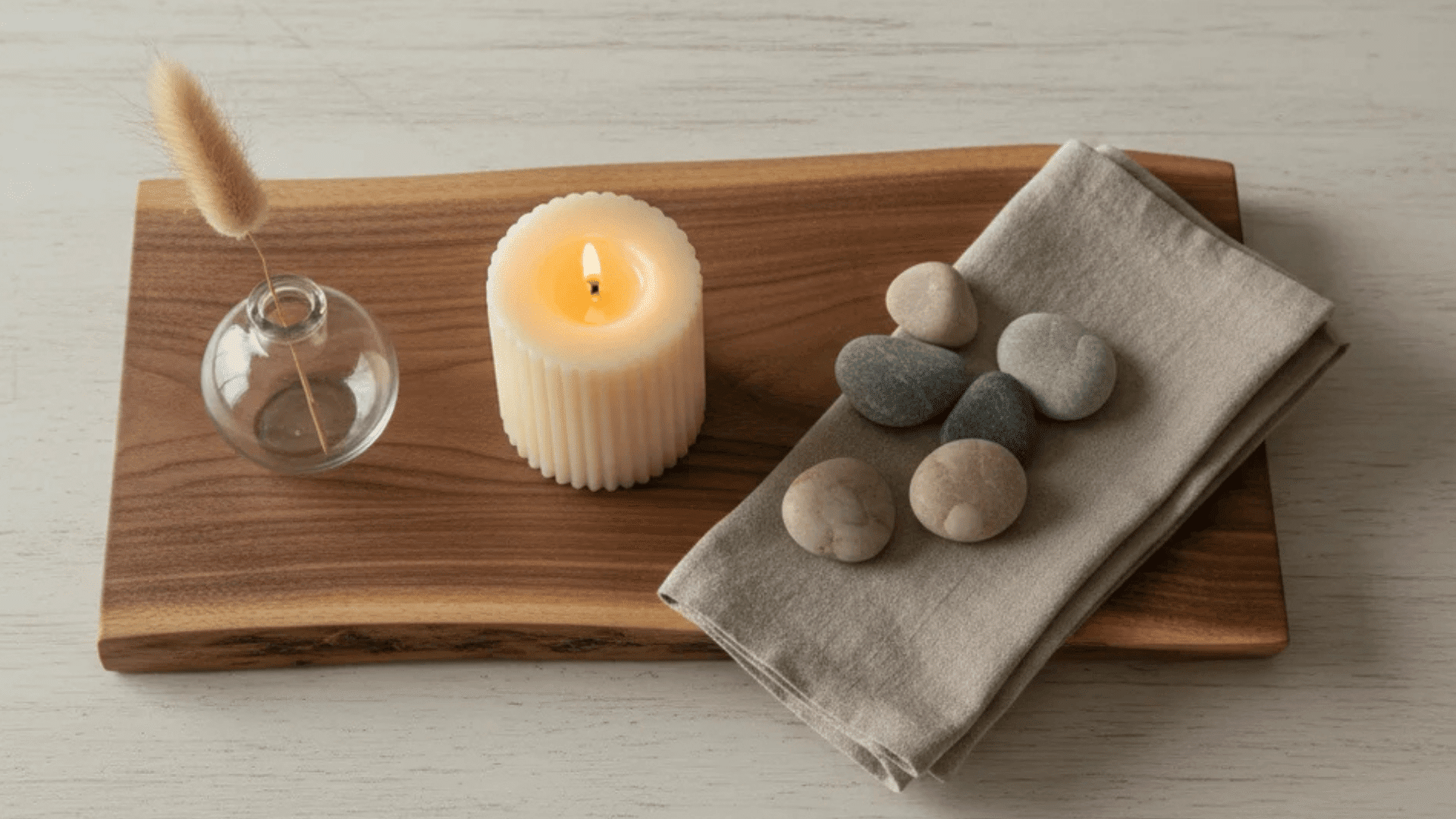a lit candle on wooden tray with small glass vase dried grass folded cloth and smooth stones arranged neatly on table