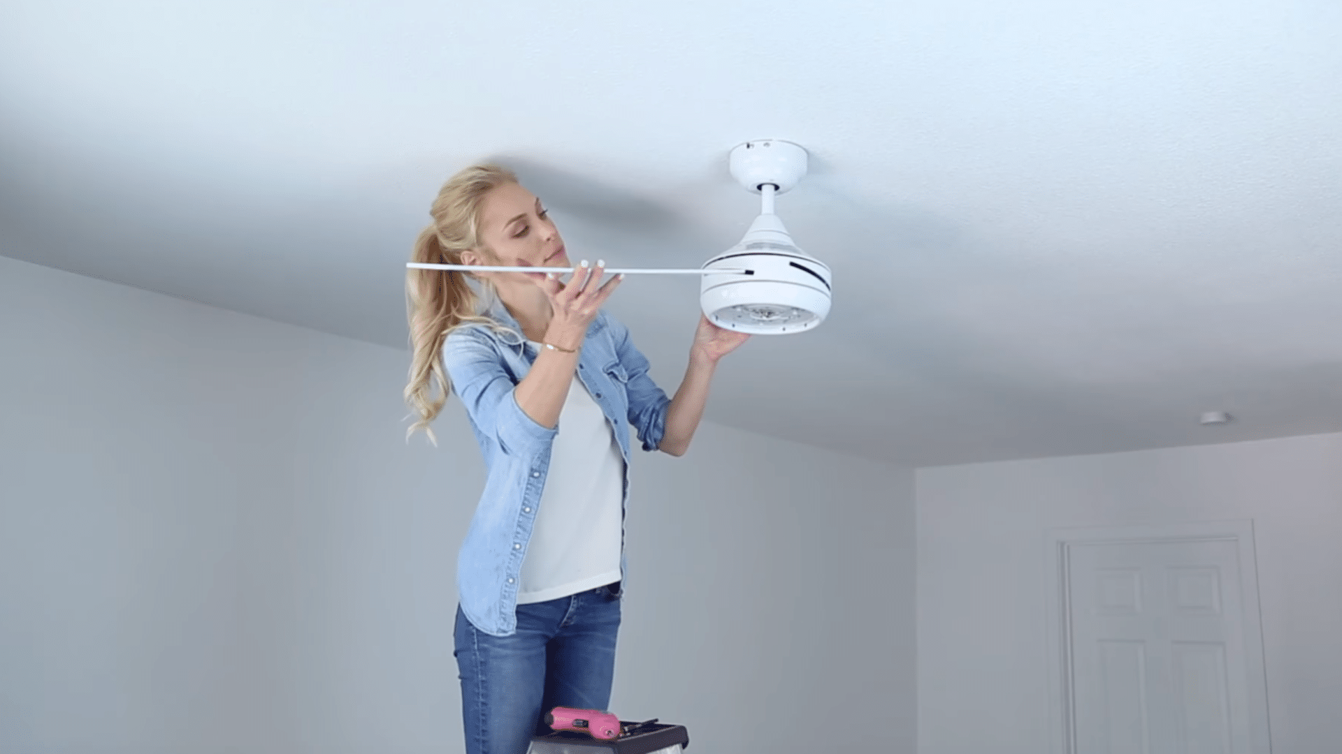 a woman standing on a ladder installing a white ceiling fan in a bright room wearing casual clothes