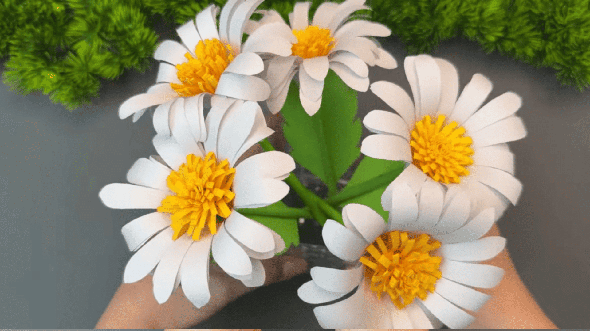 bouquet of handmade white paper daisy flowers with yellow centers and green leaves arranged in a clear glass vase on a gray table