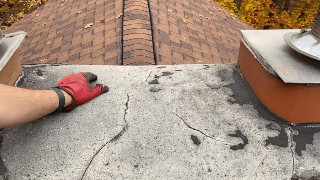 close up of cracked chimney crown with gloved hand pointing at damage on rooftop near metal chimney cap
