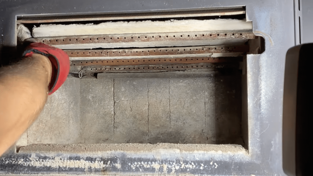 close up of stove interior with metal tubes ash buildup and gloved hand cleaning inside firebox