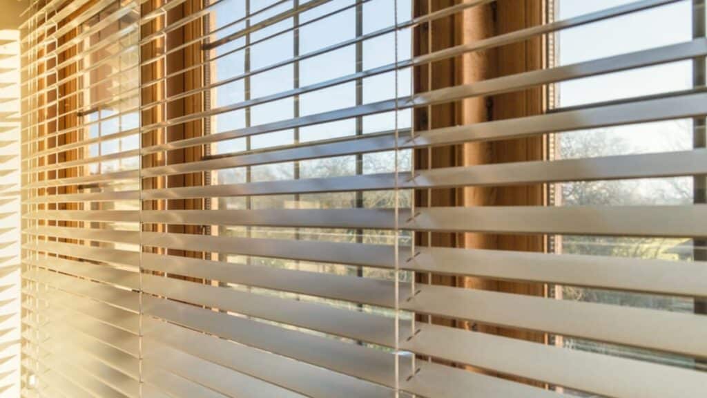 close view of white horizontal blinds on a window with sunlight streaming through slats casting soft lines across the glass