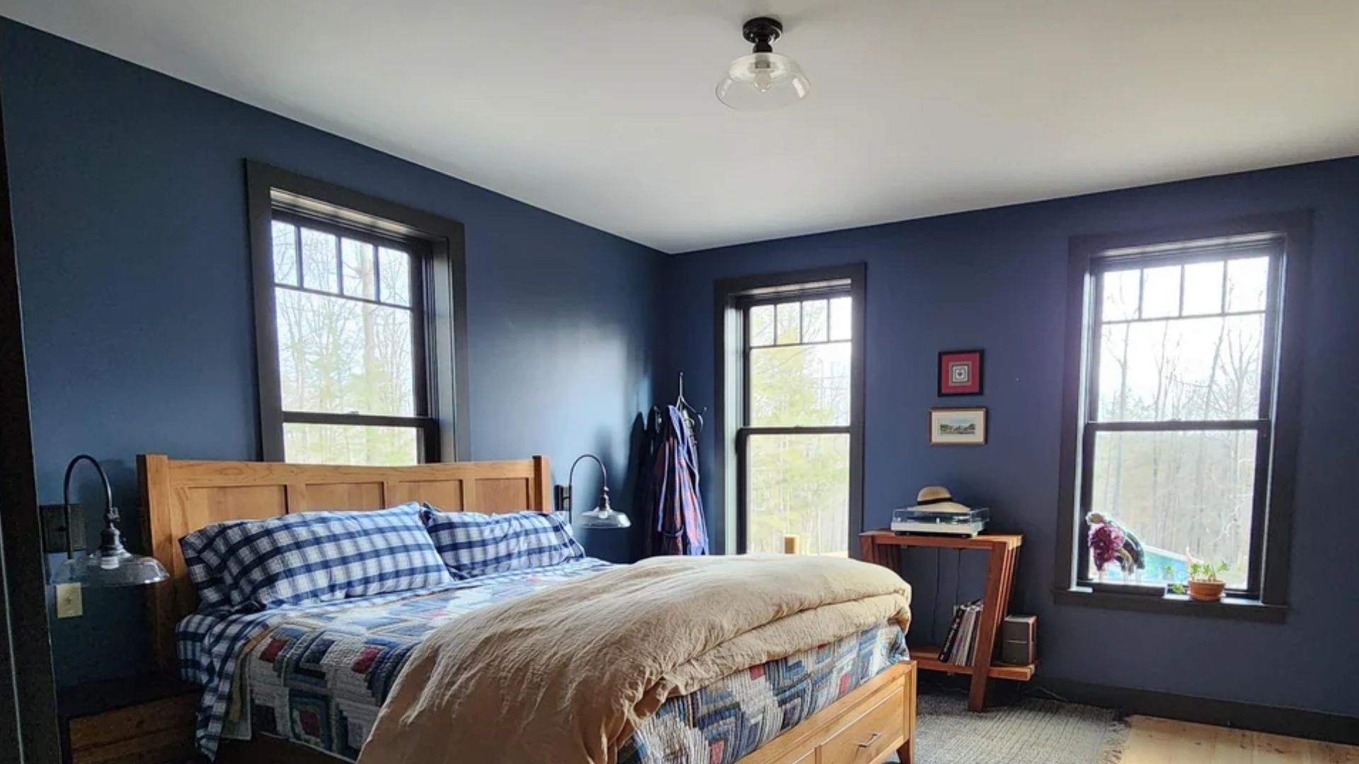 cozy bedroom with dark blue walls wooden bed with plaid bedding three windows letting in daylight and a small table with decor