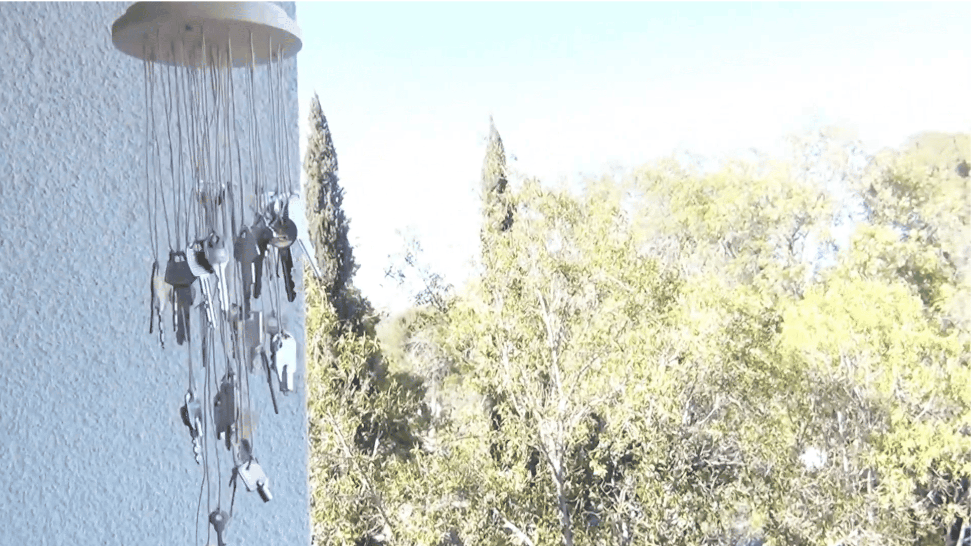 finished diy key wind chime hanging outdoors beside a wall with trees moving gently in the background