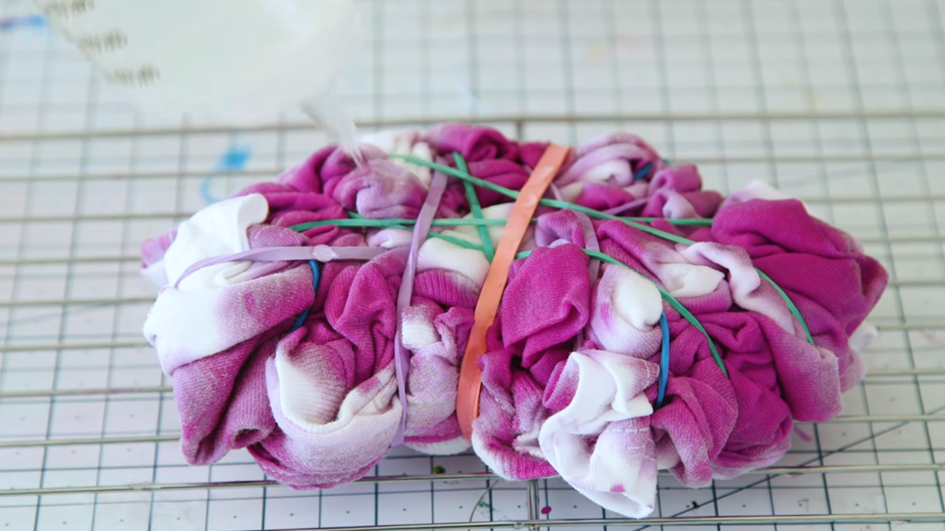 folded white tshirt dyed pink and secured with colorful rubber bands resting on a wire rack for drying