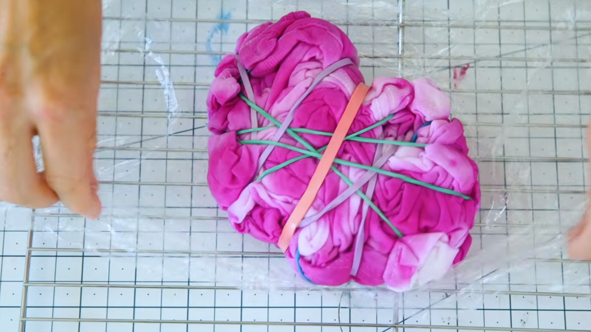 fpink tie dyed tshirt wrapped with colorful rubber bands placed on plastic wrap over a wire drying rack