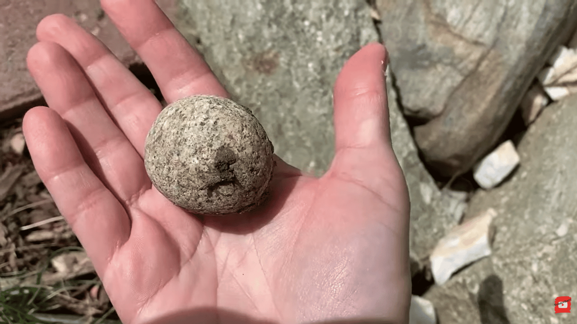 hand holding small round natural rock with rough surface outdoors with larger stones in background (1)