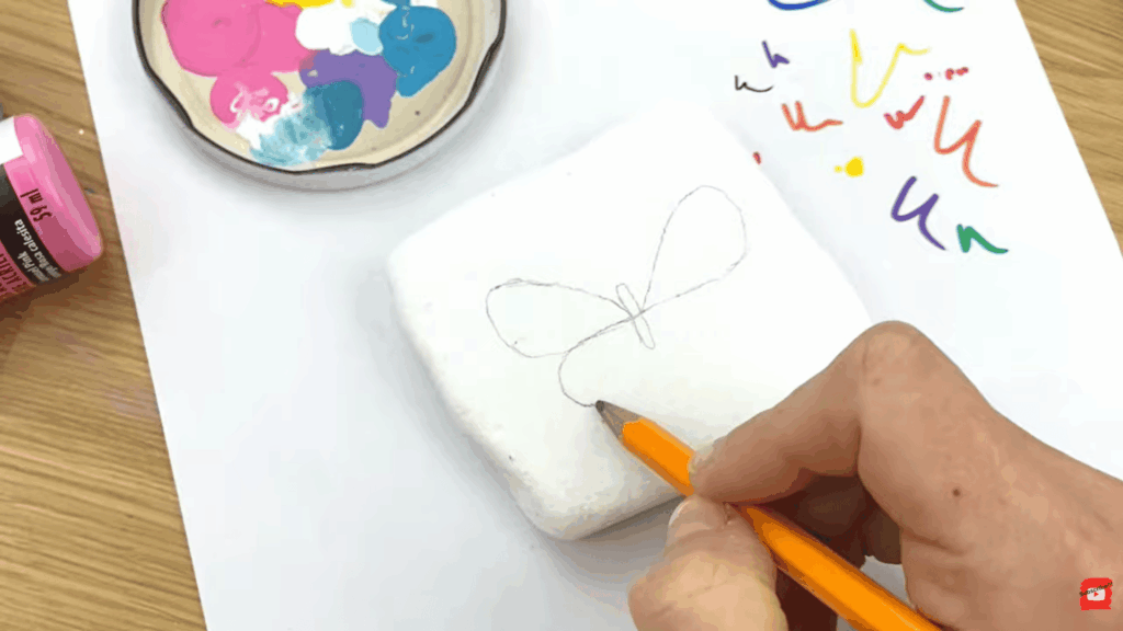 hand sketching butterfly outline on white painted rock with colorful paint palette and markers on table