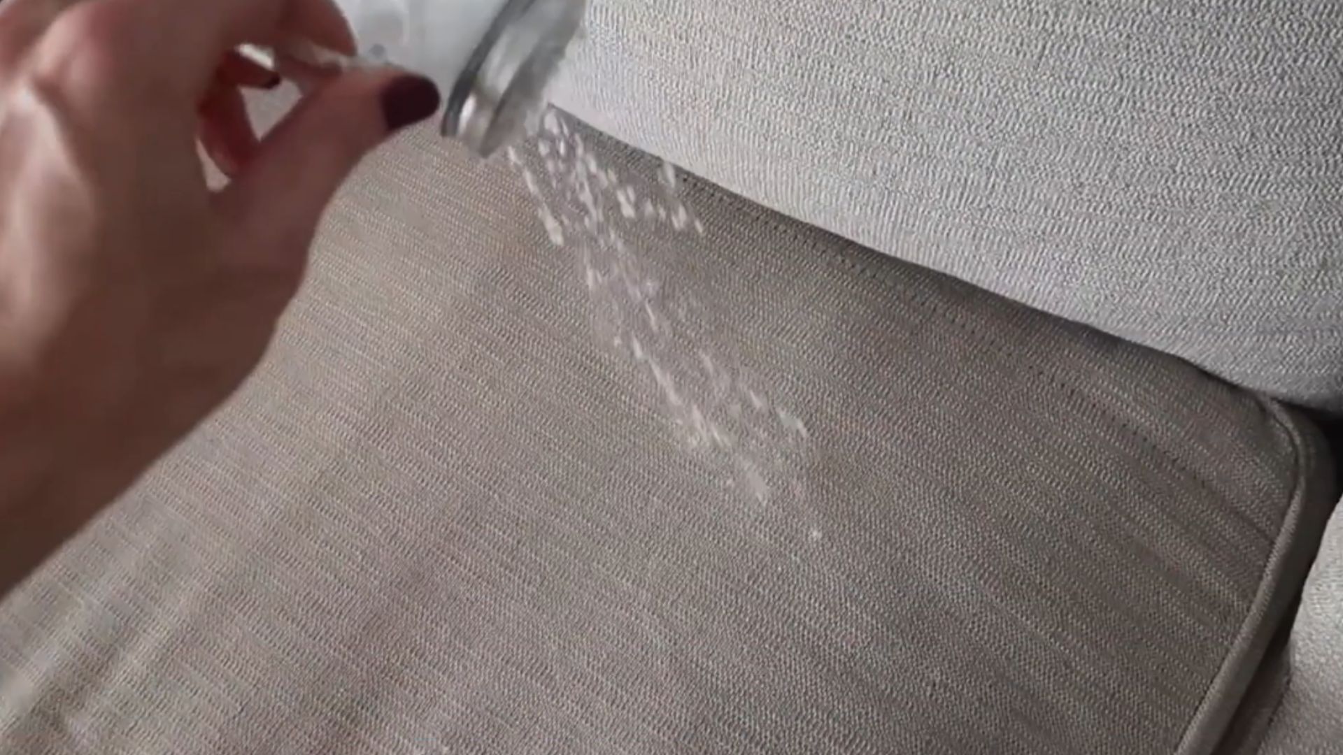 hand sprinkling white powder onto a light fabric cushion surface showing a cleaning or stain treatment process in action