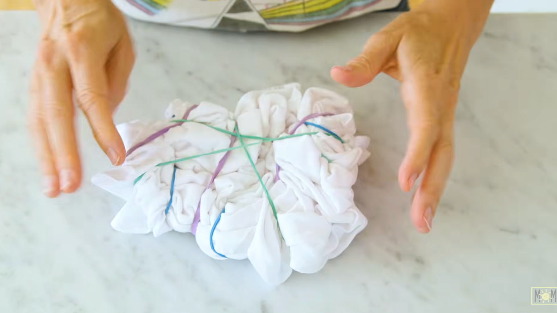 hands arranging a white folded tshirt secured with colorful rubber bands on a table for tie dye preparation