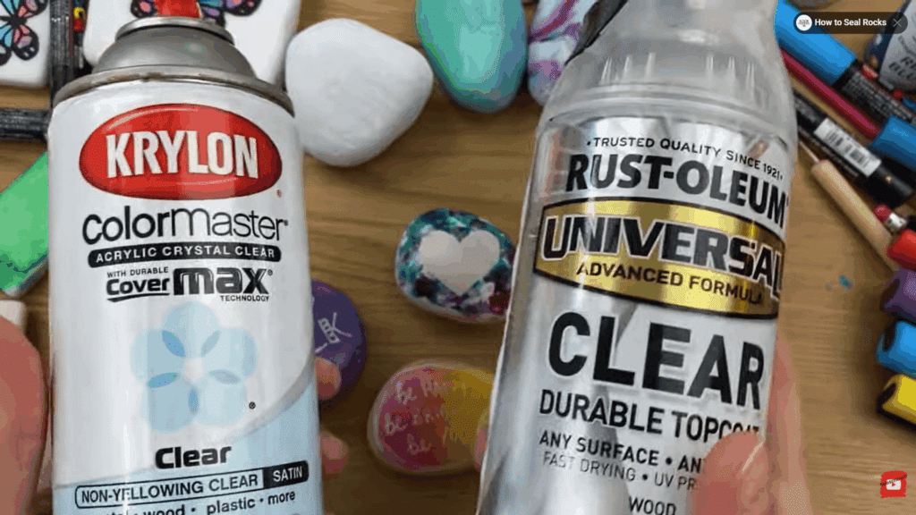 hands holding clear spray sealers for painted rocks with colorful decorated stones and art supplies on table