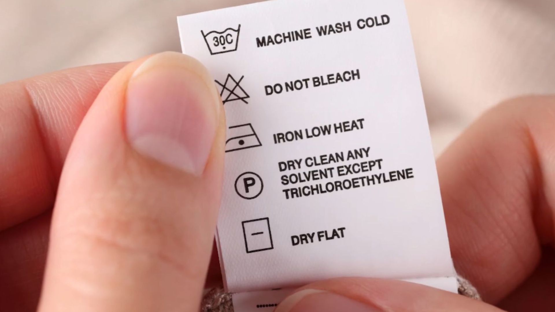 hands holding clothing care label showing washing instructions with symbols for cold wash no bleach low heat iron dry clean and dry flat