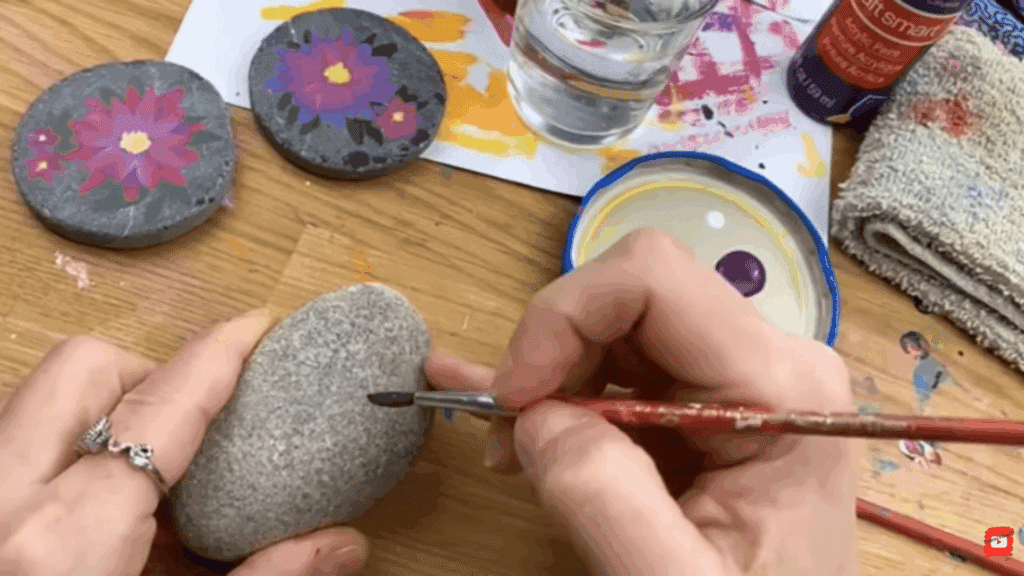 hands painting green details on smooth gray rock with floral painted stones and art supplies on wooden table