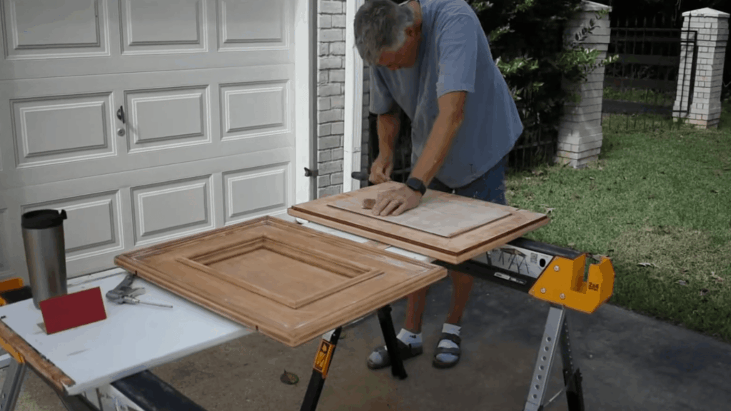 man sanding wooden cabinet door on sawhorse in driveway near garage with tools on table beside him
