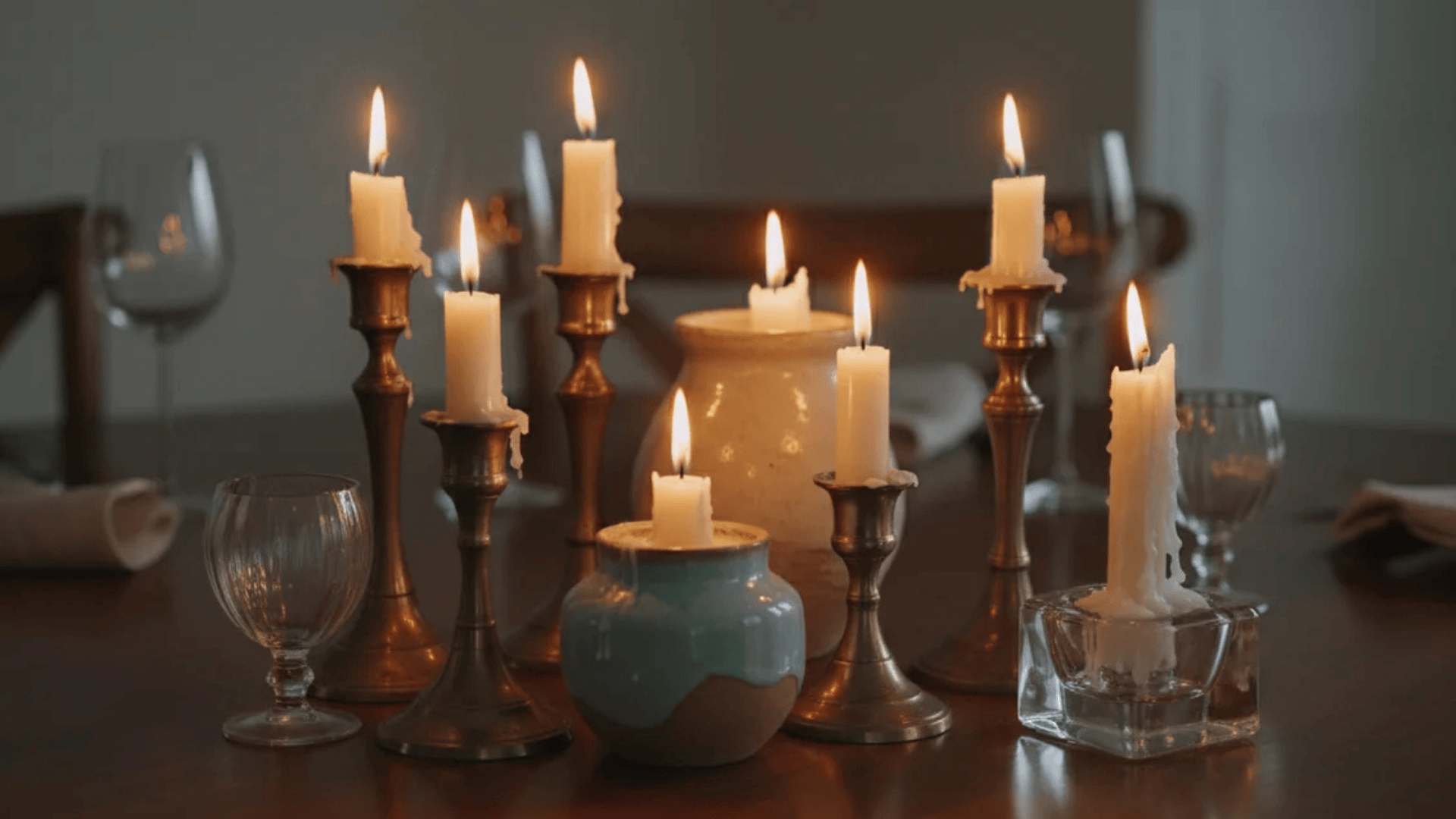 multiple lit candles in holders and small jars arranged on wooden table with glasses and plates in dim warm setting