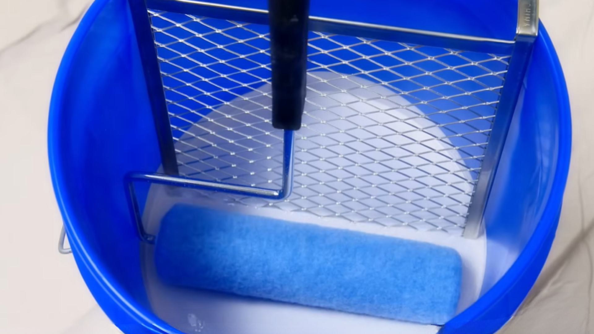 paint roller resting in blue bucket filled with white paint with metal grid used for removing excess paint before application