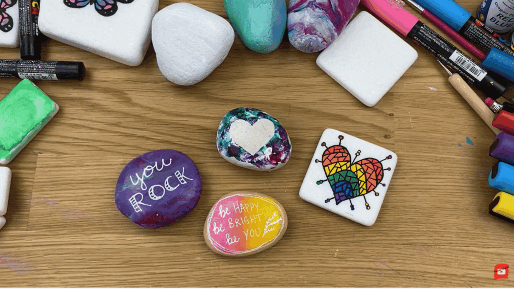 painted rocks with colorful hearts quotes and patterns on wooden table with markers and craft tools around