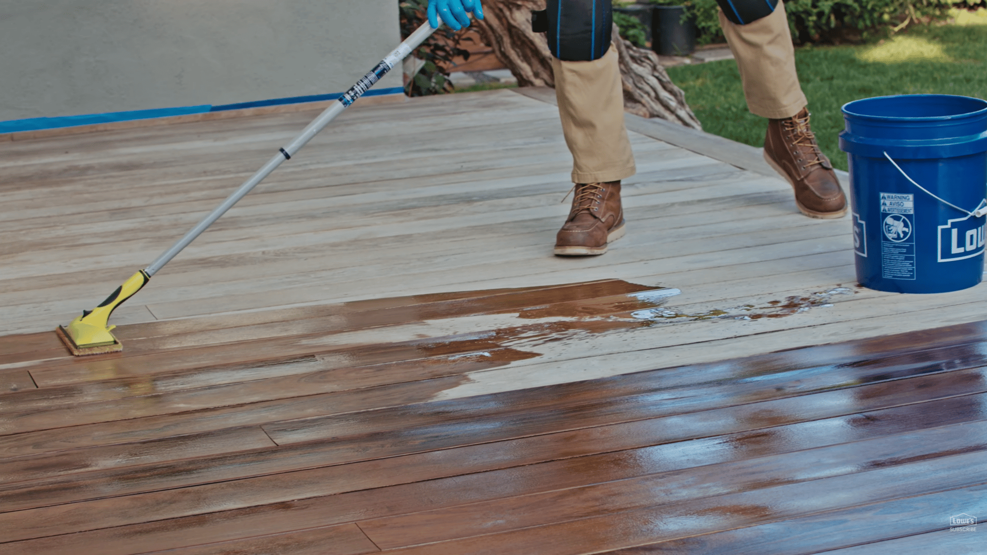 person applying dark wood stain to outdoor deck boards using long handle applicator beside blue bucket