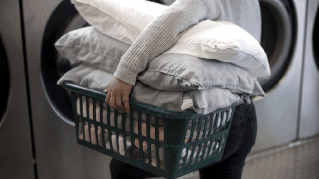 person carrying basket filled with white pillows near washing machines in a laundry room preparing to clean them