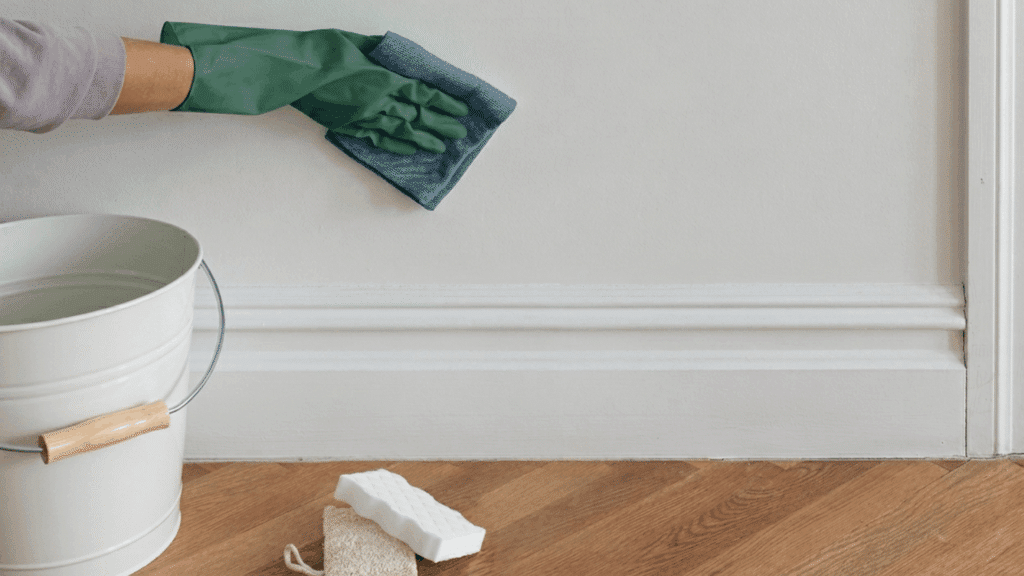 person cleaning a white wall with a cloth and gloves beside a bucket on a wooden floor