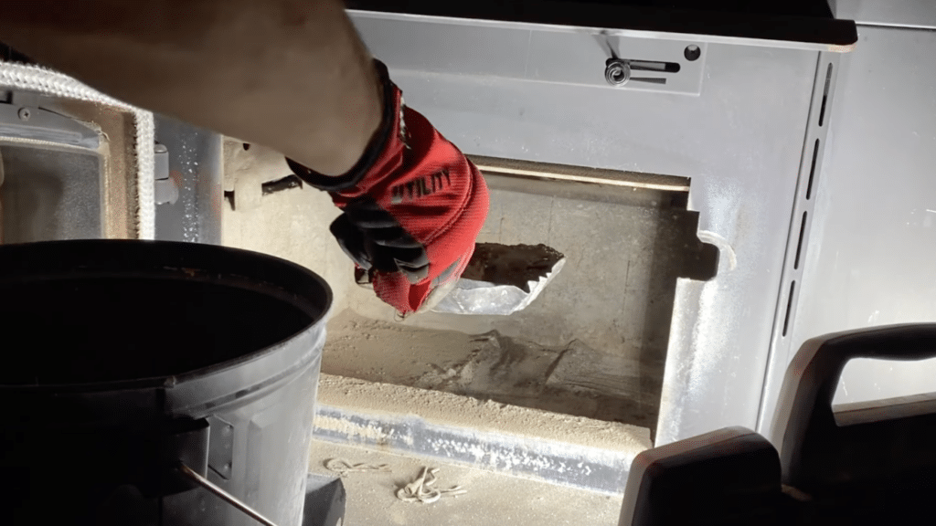 person cleaning stove interior with small shovel and bucket removing ash from firebox